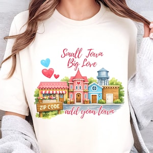 May include: Cream-colored t-shirt featuring a watercolor illustration of a small town with buildings, hearts, and the text "Small Town Big Love add your town". A sign reads "ZIP CODE".