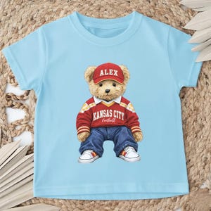 Puede incluir: Camiseta azul claro con un oso de peluche que lleva una gorra roja con "ALEX". El oso tambi&eacute;n lleva una camiseta roja y blanca de Kansas City Football, vaqueros azules y zapatillas blancas.