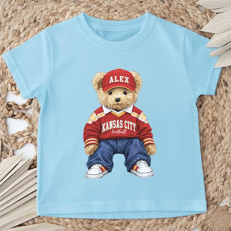 Puede incluir: Camiseta azul claro con un oso de peluche que lleva una gorra roja con "ALEX". El oso tambi&eacute;n lleva una camiseta roja y blanca de Kansas City Football, vaqueros azules y zapatillas blancas.