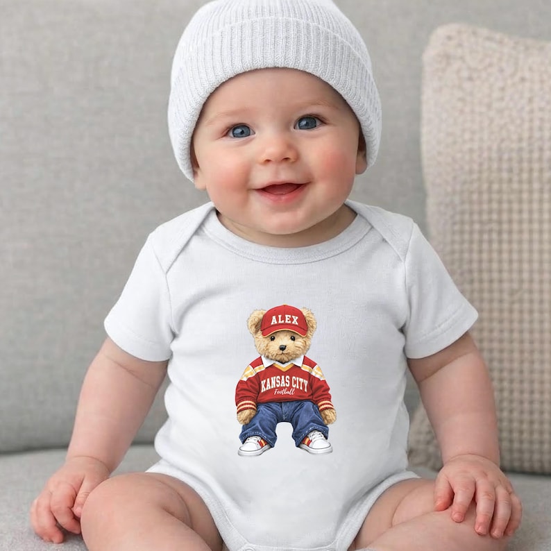 Puede incluir: Un beb&eacute; sonriente con un body blanco y un gorro blanco. El body presenta un oso de peluche con una gorra roja con "ALEX", un su&eacute;ter rojo y amarillo, vaqueros azules y zapatillas blancas. El texto "Kansas City Football" est&aacute; en el su&eacute;ter.