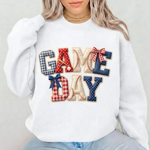 以下が含まれることがあります： 「GAME DAY」の文字が入った白いスウェットシャツ。文字は野球のステッチ、バット、赤、白、青の模様のリボンで装飾されています。野球ボールも含まれています。
