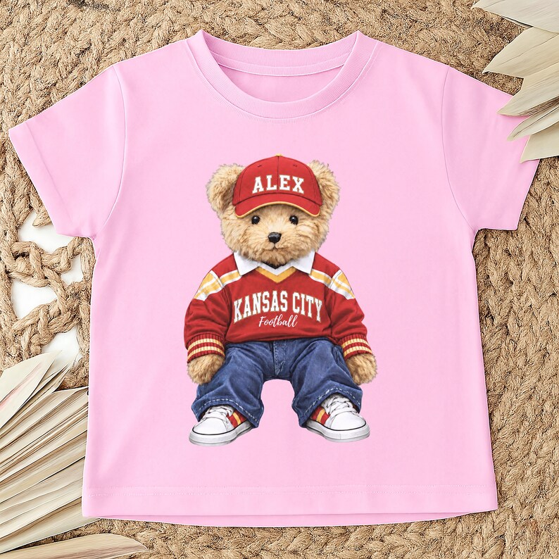 Puede incluir: Camiseta rosa con un oso de peluche que lleva una gorra roja con "ALEX", un su&eacute;ter rojo y amarillo de Kansas City Football, vaqueros azules y zapatillas blancas. El oso de peluche es el motivo principal de la camiseta.