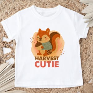 以下が含まれることがあります： 白いTシャツ。どんぐりを持った漫画のリス、緑のセーター、茶色とオレンジ色の尻尾が特徴です。「HARVEST CUTIE」の文字が赤とオレンジ色でプリントされています。このシャツはおそらく子供用です。