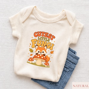 Pode incluir: Um body de manga curta na cor creme com um gráfico de uma raposa sentada num cogumelo vermelho com pintas brancas. O texto "CUTEST LITTLE FUNGI" está em letras laranja e castanhas, rodeado por pequenas folhas e estrelas, com ganga azul.