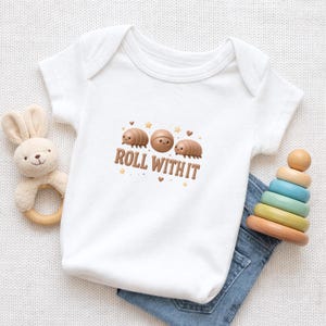 Może przedstawiać: Białe body dla dziecka z grafiką trzech kreskówkowych stonóg i brązowym napisem "ROLL WITH IT". Body połączone jest z niebieskimi dżinsami, drewnianą zabawką do układania i pluszową zabawką w kształcie królika.