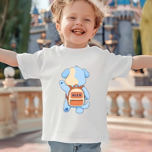 Op de afbeelding: Een wit t-shirt met korte mouwen en een afbeelding van een lichtblauwe puppy met een crèmekleurige vlek op de kop. De puppy is van achteren afgebeeld en draagt een oranje rugzak met de naam ALEX in hoofdletters op de voorzijde.