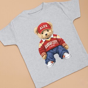 Puede incluir: Camiseta gris claro con un oso de peluche que lleva una gorra roja con "ALEX", un su&eacute;ter rojo y blanco con "KANSAS CITY Football" y vaqueros azules. Zapatillas blancas con detalles rojos completan el conjunto.