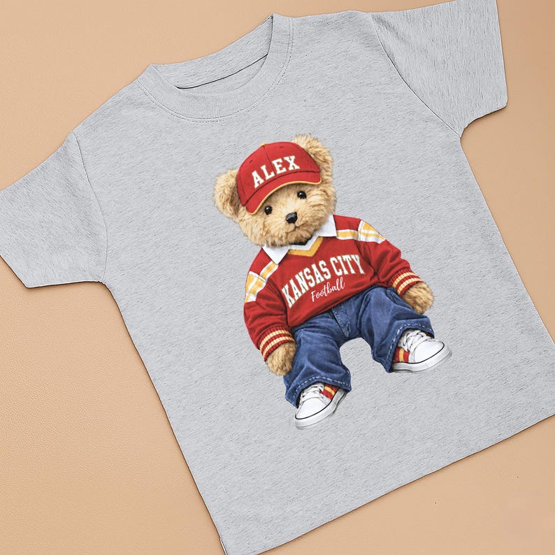 Puede incluir: Camiseta gris claro con un oso de peluche que lleva una gorra roja con "ALEX", un su&eacute;ter rojo y blanco con "KANSAS CITY Football" y vaqueros azules. Zapatillas blancas con detalles rojos completan el conjunto.