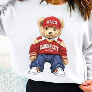 Puede incluir: Una sudadera blanca con un oso de peluche que lleva una camiseta de f&uacute;tbol roja de Kansas City, vaqueros azules, una gorra roja con "ALEX" y zapatillas blancas. El oso est&aacute; sentado. La sudadera es de cuello redondo.