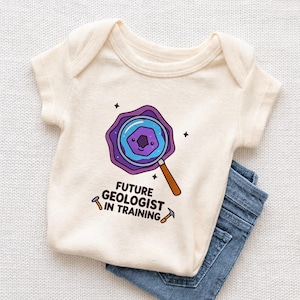 May include: Cream-colored baby onesie with the text "FUTURE GEOLOGIST IN TRAINING" and a graphic of a magnifying glass over a geological formation. The onesie is paired with blue denim jeans.