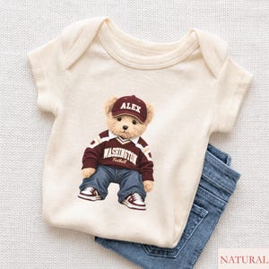 May include: A cream-coloured short-sleeve baby bodysuit featuring a graphic of a teddy bear wearing a maroon baseball cap with the name ALEX, a maroon V-neck jumper with WASHINGTON Football printed on the front, blue jeans, and trainers.
