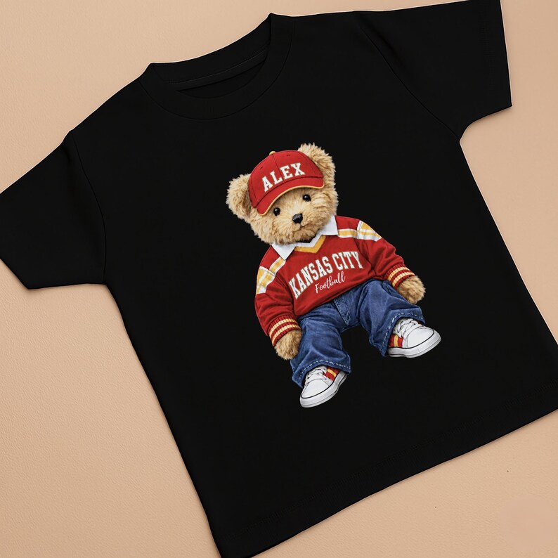 Puede incluir: Camiseta negra con un oso de peluche que lleva una gorra roja con "ALEX", un su&eacute;ter rojo y amarillo de Kansas City Football, vaqueros azules y zapatillas blancas. El oso de peluche es el gr&aacute;fico central de la camiseta.