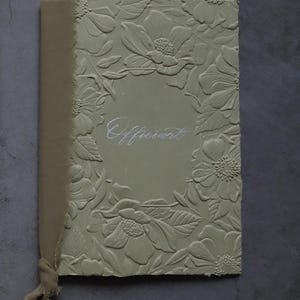 May include: A sage green book with embossed floral designs and the word "Officiant" in silver script. The book has a tan fabric spine and a matching ribbon tie.
