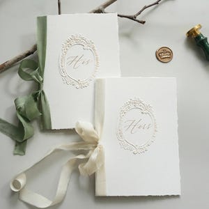 Op de afbeelding: Twee witte, getextureerde notitieboekjes met de woorden "His" en "Hers" in elegant schrift in sierlijke kaders. Elk boek is gebonden met een zijden lint in groen- en crèmetinten. Een gouden zegel en een stempel met groene handgreep zijn ook aanwezig.