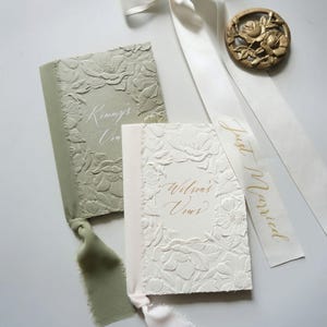 Peut inclure: Deux livrets de vœux de mariage avec des couvertures gaufrées à motifs floraux. L'un est vert sauge, l'autre est blanc. Le livre blanc porte l'inscription "Wilson's Vows" en lettres dorées. Un ruban blanc avec "Just Married" en lettres dorées est également présent.