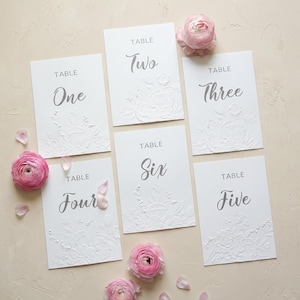May include: Six white table cards with embossed floral designs and gray text. Each card is labeled with a table number: One, Two, Three, Four, Five, and Six. Pink flowers and petals are scattered around the cards.