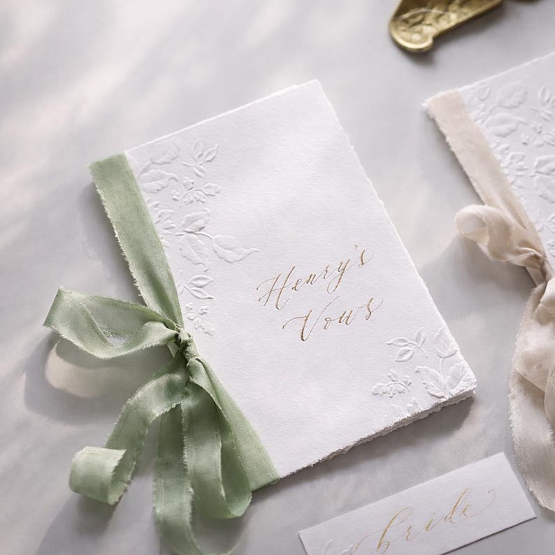 May include: White wedding vow booklet with embossed floral design and sage green ribbon. The text "Henry's Vows" is written in gold script. A small card with the word "Bride" is also visible.