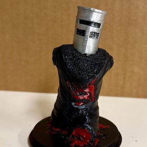 Può includere: Una statuina stampata in 3D del Cavaliere Nero di Monty Python e il Sacro Graal. Il cavaliere è raffigurato senza testa, con un elmo argentato e un'armatura nera con dettagli rossi, su una base nera.