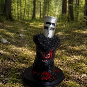 May include: A figurine of a knight wearing a silver cylindrical helmet with a horizontal visor slit, set atop a black torso piece with textured black shoulders and a red painted emblem on the chest, all mounted on a circular black base.