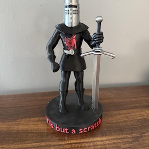 May include: A black figurine of a knight in armor, holding a sword. The knight has a silver helmet and a red emblem on the chest. The base reads "Tis but a scratch" in red lettering.