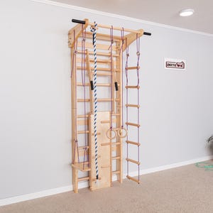 May include: Wooden wall-mounted climbing frame with multiple features. Includes a ladder, rope, rings, and a climbing rope with blue and white stripes. The frame is made of light-colored wood and is designed for indoor use.