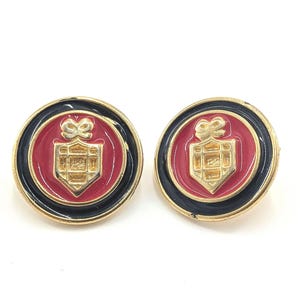 May include: A pair of round earrings with a gold-tone border and a black enamel ring. The center features a red enamel background with a gold-tone shield design and a bow at the top. The earrings are set against a white background.