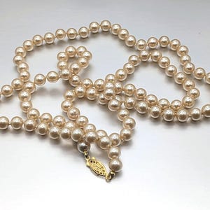 May include: A strand of cream-colored faux pearls. The necklace is composed of numerous round beads, strung together. A gold-colored clasp secures the necklace. The pearls are evenly sized and have a subtle luster.