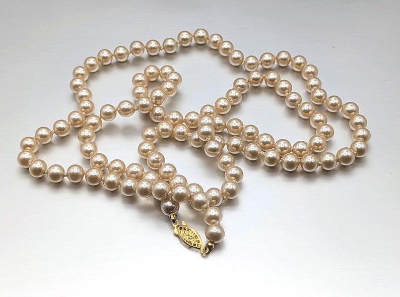 May include: A strand of cream-colored faux pearls. The necklace is composed of numerous round beads, strung together. A gold-colored clasp secures the necklace. The pearls are evenly sized and have a subtle luster.
