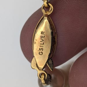 May include: Close-up of a gold-toned clasp with the engraved text "GSILVER". The clasp is connected to a strand of light-colored, round beads. The clasp is a teardrop shape with a small loop at each end.