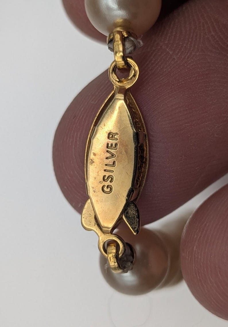 May include: Close-up of a gold-toned clasp with the engraved text "GSILVER". The clasp is connected to a strand of light-colored, round beads. The clasp is a teardrop shape with a small loop at each end.