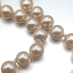 May include: Close-up of a strand of faux pearls. The pearls are a creamy, off-white color and are strung together with a matching thread. The pearls have a glossy finish and reflect light.