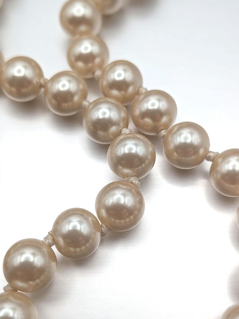 May include: Close-up of a strand of faux pearls. The pearls are a creamy, off-white color and are strung together with a matching thread. The pearls have a glossy finish and reflect light.