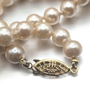 May include: A close-up of a cream-colored pearl necklace with a gold-toned clasp. The pearls are round and evenly sized, strung together with a delicate thread. The clasp is ornate with a decorative design.