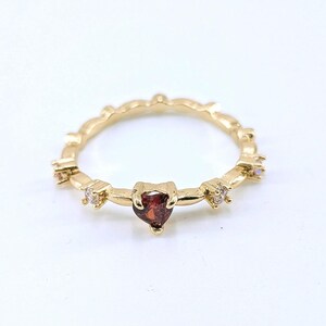 May include: A gold-colored ring with a heart-shaped, dark red gemstone at the center. The ring features small, clear gemstones and a delicate, scalloped band. The ring is set against a white background.