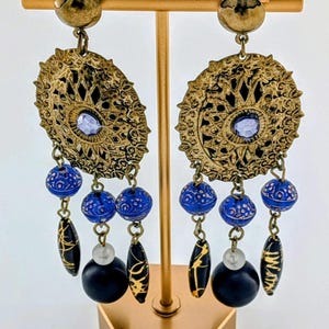 May include: Ornate gold-tone earrings with intricate filigree design. Each earring features a central lavender-colored stone, dangling blue and black beads, and black teardrop-shaped accents with gold detailing. The earrings are displayed on a gold stand.