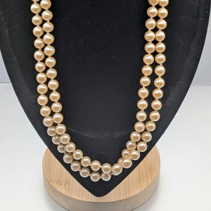 May include: A double-strand necklace with faux pearls. The necklace is displayed on a black velvet bust with a wooden base. The pearls are a warm cream color, and the necklace is draped in a classic style.