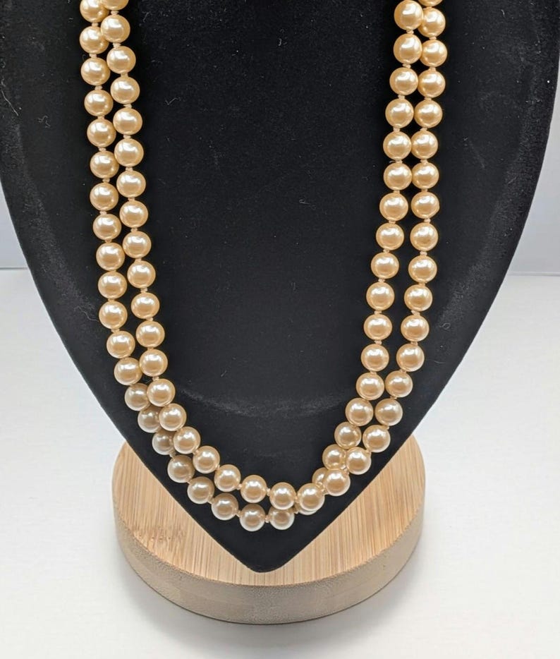 May include: A double-strand necklace with faux pearls. The necklace is displayed on a black velvet bust with a wooden base. The pearls are a warm cream color, and the necklace is draped in a classic style.