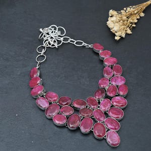 May include: A statement necklace featuring multiple oval-shaped, faceted deep pink gemstones set in silver-toned metal bezels. The stones are arranged in a tiered, cascading pattern, connected by a silver-toned chain with circular links at the top.