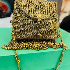Può includere: Una borsa a mano in metallo intrecciato color oro con tracolla a catena e frange di perline. La borsa ha una forma rettangolare con una patta superiore curva fissata da una chiusura.