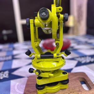 May include: A bright yellow theodolite, a surveying instrument, is displayed on a wooden cutting board. The theodolite has multiple adjustment knobs and a sighting scope. The instrument is used for measuring angles in the horizontal and vertical planes.