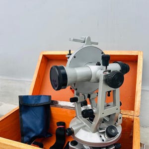 May include: A silver theodolite, a surveying instrument, is displayed inside an open wooden box. The theodolite has black knobs and lenses. A blue pouch and black accessories are also in the box. The box is made of light brown wood.