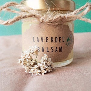 Könnte beinhalten: Ein kleines Glas mit goldenem Deckel, mit Bindfaden gebunden und einem Etikett mit der Aufschrift "LAVENDEL BALSAM". Das Glas ist von getrockneten Blumen umgeben. Der Hintergrund ist ein zartes Rosa und Türkis.