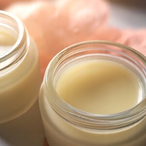 Könnte beinhalten: Zwei kleine, klare Glasdosen gefüllt mit einer glatten, blassgelben Creme. Die Dosen haben weite, offene Öffnungen, die die cremige, undurchsichtige Substanz im Inneren zeigen. Die Behälter stehen nebeneinander und zeigen ihr einfaches, zylindrisches Design.