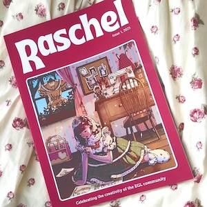 以下が含まれることがあります： 表紙がマルーン色の雑誌「Raschel」で、緑色のドレスを着た女の子のイラストが描かれています。雑誌は花柄の生地の上にあります。「Issue 1, 2025」の文字が見えます。EGLコミュニティの創造性を祝っています。