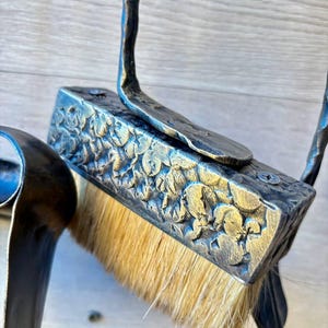 May include: Close-up of a handcrafted, ornate metal hearth brush. The brush features a rectangular head with a floral pattern and natural bristles. The handle is curved and appears to be made of the same dark metal. The brush is designed for cleaning a fireplace.