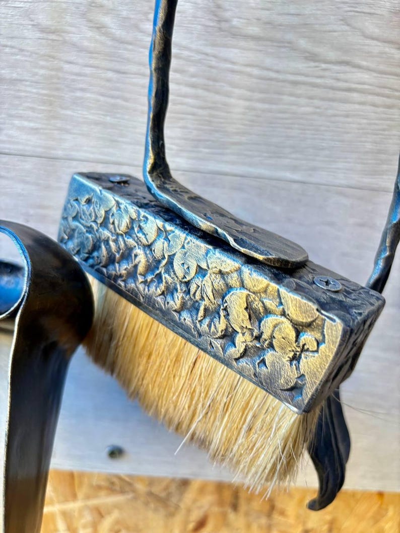 May include: Close-up of a handcrafted, ornate metal hearth brush. The brush features a rectangular head with a floral pattern and natural bristles. The handle is curved and appears to be made of the same dark metal. The brush is designed for cleaning a fireplace.