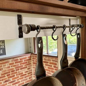 May include: A rustic wrought iron pot rack with four black frying pans suspended from it. The rack is fixed beneath a wooden shelf and has curved hooks. The pans have long handles. A brick wall and window are in the background.