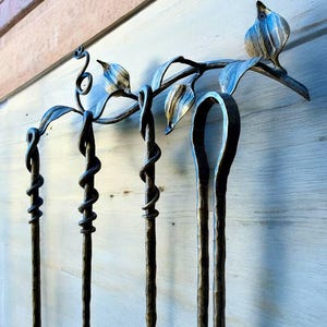 May include: A handcrafted, wrought iron fireplace tool set. The set includes a stand with a bird and leaf design, and four tools with twisted and looped details. The metal has a dark, aged finish, and the tools are designed to hang from the stand.