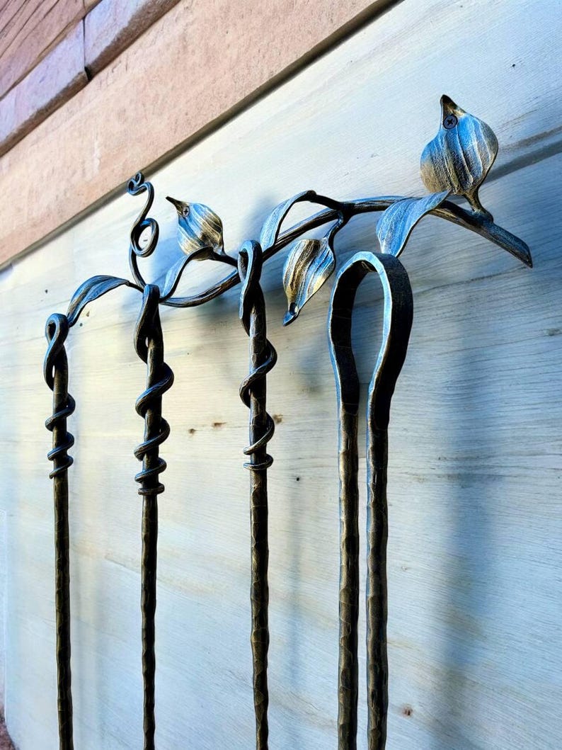 May include: A handcrafted, wrought iron fireplace tool set. The set includes a stand with a bird and leaf design, and four tools with twisted and looped details. The metal has a dark, aged finish, and the tools are designed to hang from the stand.