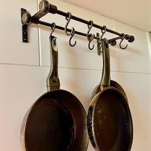 May include: A dark metal pot rack with several curved hooks, holding two black frying pans. The rack is mounted on a white tiled wall. The pans have dark, well-used interiors, suggesting frequent use in cooking.